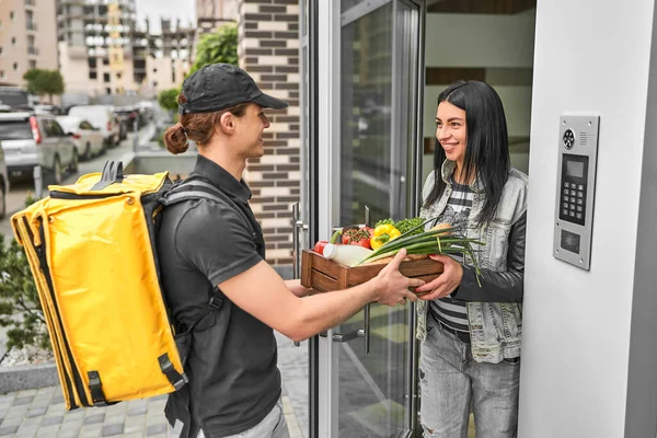 提着背包和食物的快递员在网上向顾客的家里下了订单 那女人在她家门口接蔬菜 — 图库照片