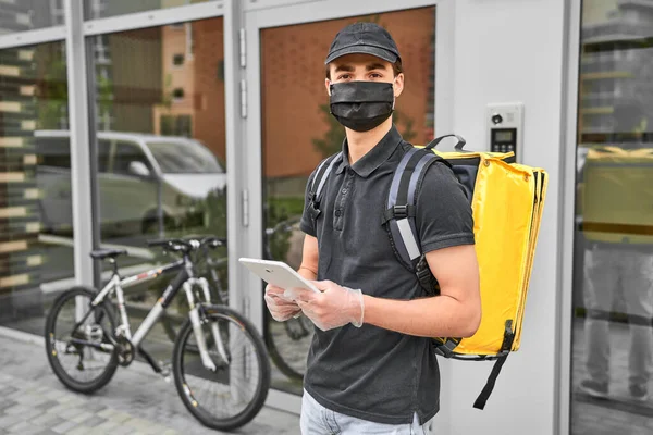 Delivery man in protective mask delivered an online order on the bicycle. He have yellow backpack and tablet. On the city street.
