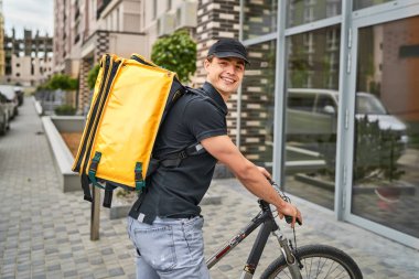 Modern bir evin yanında bisikletli, sarı sırt çantalı, mutlu bir teslimatçı..
