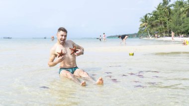 Seyahat oğlan kırmızı starfishes yapar komik fotoğraflar Phu Quoc Adası kristal berraklığında su ile denizde oturuyor