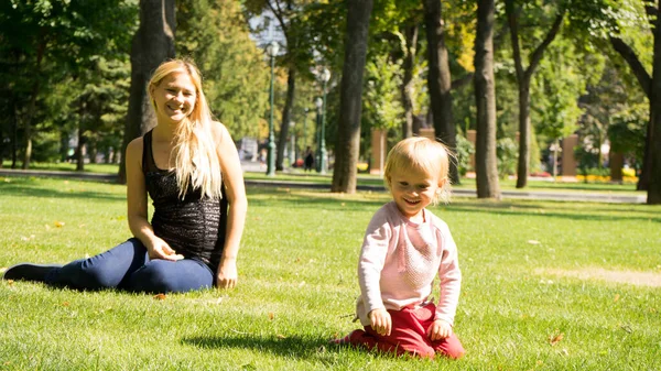 Beautiful mother and daughter with blonde hair playing together in the sunny park .Mother and daughter sitting together on green grass.Mother and daughter having funny conversation.Happy family concep