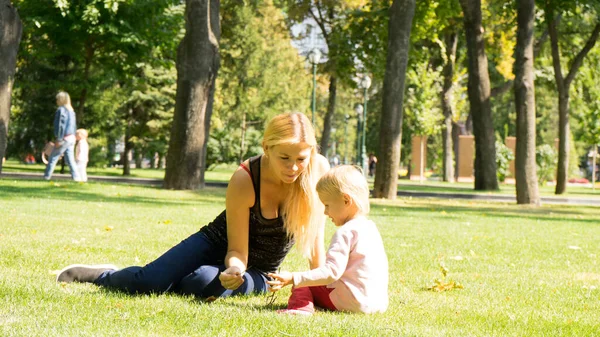 Beautiful mother and daughter with blonde hair playing together in the sunny park .Mother and daughter sitting together on green grass.Mother and daughter having funny conversation.Happy family concep