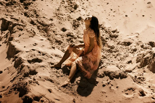 穿着粉红色连衣裙的性感女士坐在沙漠里闭着眼睛的沙子上 — 图库照片
