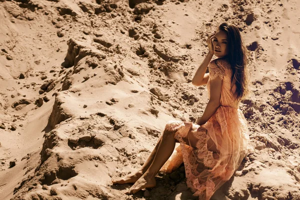 魅力的な少女ピンクのドレスを着て砂の上に座っているし 砂漠でカメラを見て — ストック写真