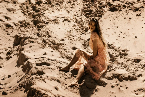 穿着粉红色连衣裙的漂亮女孩坐在沙滩上 看着沙漠里的照相机 — 图库照片