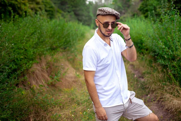 Portret van Prachtige jongeman model in hoed en zonnebril weg te kijken op het groene park — Stockfoto