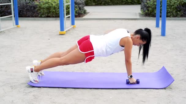 Hübsche Frau trainiert ihre Bizeps-Muskeln mit Hanteln auf einem lila Karemat an einem warmen Sommertag im Park — Stockvideo
