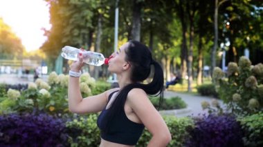 Güzel esmer sporcu kadın parkta pratik yaptıktan sonra su içiyor.