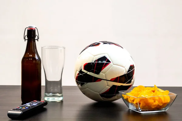 People prepared to watch football on TV with beer. There\'s beer on the table, ball, TV remote, snacks. Craft beer. Light background.