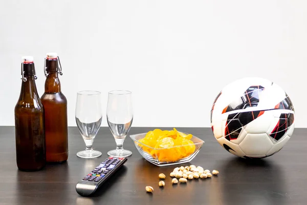 People prepared to watch football on TV with beer. There\'s beer on the table, ball, TV remote, snacks. Craft beer. Light background.
