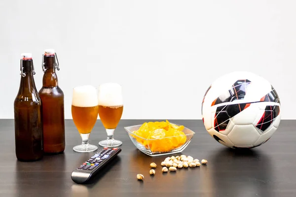 People prepared to watch football on TV with beer. There\'s beer on the table, ball, TV remote, snacks. Craft beer. Light background.