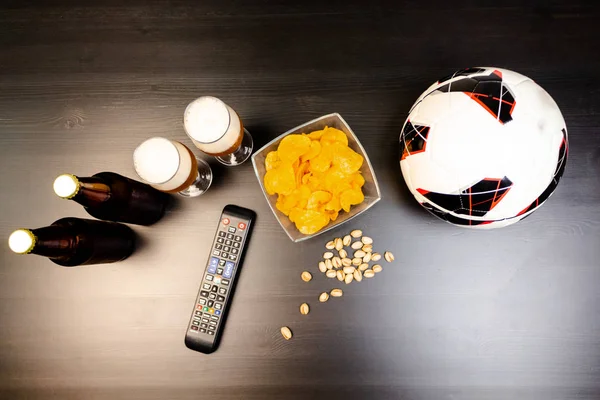 People prepared to watch football on TV with beer. There\'s beer on the table, ball, TV remote, snacks. Craft beer. Light background. The view from the top.