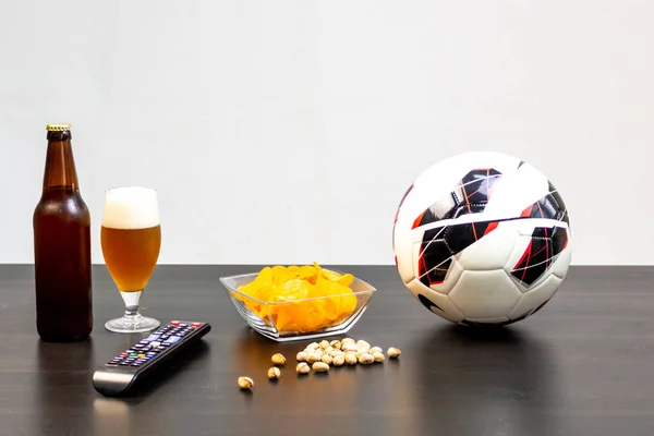 People prepared to watch football on TV with beer. There\'s beer on the table, ball, TV remote, snacks. Craft beer. Light background.