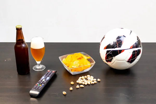 People prepared to watch football on TV with beer. There's beer on the table, ball, TV remote, snacks. Craft beer. Light background.