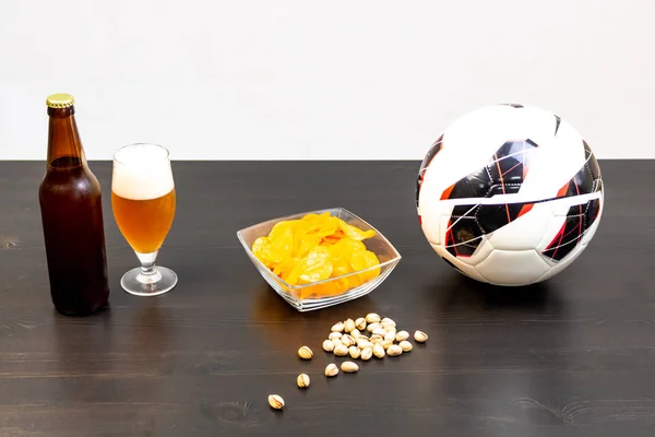 People prepared to watch football on TV with beer. There\'s beer on the table, ball, TV remote, snacks. Craft beer. Light background.