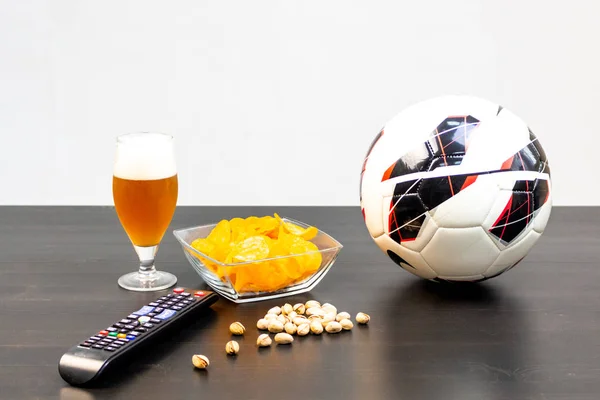 People prepared to watch football on TV with beer. There\'s beer on the table, ball, TV remote, snacks. Craft beer. Light background.