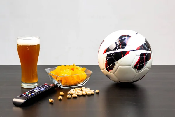 People prepared to watch football on TV with beer. There\'s beer on the table, ball, TV remote, snacks. Craft beer. Light background.