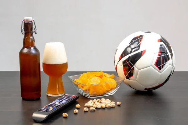 People prepared to watch football on TV with beer. There\'s beer on the table, ball, TV remote, snacks. Craft beer. Light background.