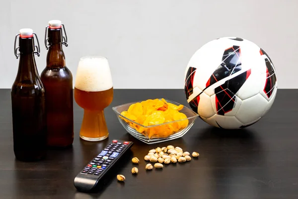 People prepared to watch football on TV with beer. There\'s beer on the table, ball, TV remote, snacks. Craft beer. Light background.