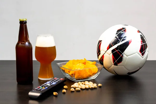 People prepared to watch football on TV with beer. There\'s beer on the table, ball, TV remote, snacks. Craft beer. Light background.