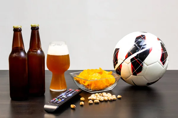 People prepared to watch football on TV with beer. There\'s beer on the table, ball, TV remote, snacks. Craft beer. Light background.