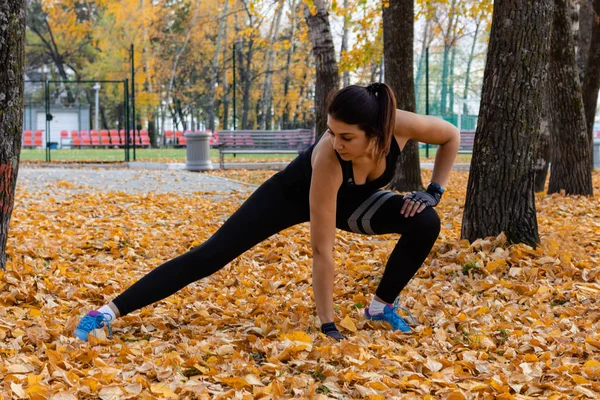 Khabarovsk, Russia - 07 ottobre 2018: Una donna attraente in abiti sportivi che fa esercizi sportivi in natura contro il tramonto e il fiume Amur, ama la ginnastica, impasta le gambe. Giovane ragazza attiva — Foto Stock