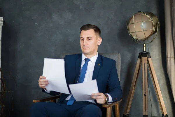 Confident man lawyer in formal wear reading paper documents while sitting in modern office space interior, young handsome male economist studying dossier files before an important meeting with partner