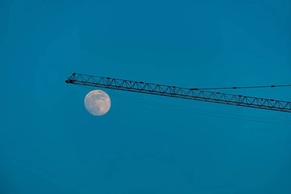 Grúa en un sitio de construcción contra el cielo azul y una luna —  Fotos de Stock