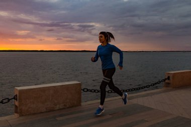 Khabarovsk, Russia - Oct 07, 2018: Young and attractive girl running at sunset in the city Park. Healthy fitness woman running outdoors. Amur river in Khabarovsk horizon in the background. clipart