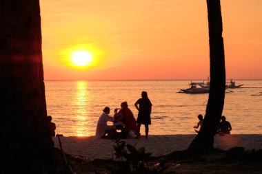 Boracay, Filipinler - 27 Ocak 2020 Boracay Adası 'nda gün batımı. Yelkencilik ve turistlerle dolu diğer geleneksel tekneler batan güneşin arka planına karşı. Turistler sahilde oturuyor ve palmiye ağaçlarının arasında manzaraya bakıyor.