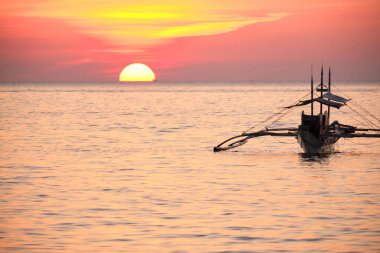 Boracay, Filipinler - 27 Ocak 2020 Boracay Adası 'nda gün batımı. Yelkencilik ve turistlerle dolu diğer geleneksel tekneler batan güneşin arka planına karşı. Turistler sahilde oturuyor ve palmiye ağaçlarının arasında manzaraya bakıyor.