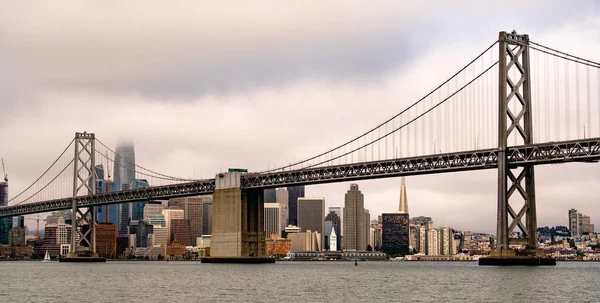 The city is shrouded in fog and the Bay Bridge stands framing the waterfront and skyline of San Francisco