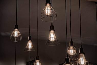 Siyah kablolar üzerinde tavandan asılı metal tel abajurlar. Cam ampuller içinde parlayan filamentler ile Lambalar. Çeşitli karkas tel şekillerin geometrisi. Koyu arka planlı parlayan avizeler. Farklı boyutlarda geometrik tel karkas avizeler.