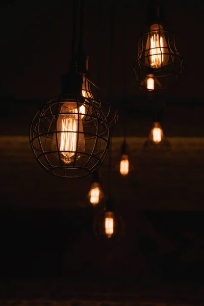 Industrial style lanterns hanged from ceiling. Metal wire lampshades in darkness. Lamps with glowing filaments inside Edison glass light bulbs. Urban style interior lighting. Wire cage lamps closeup. Geometric wire carcass chandeliers.