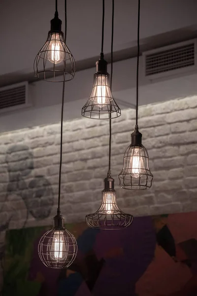 Black wire lamps hanged on black cable from ceiling on old brick wall background. Glowing filaments inside glass lightbulbs. Geometric carcass shapes. Urban style interior lighting concept. Wire chandeliers closeup. Edison retro illumination