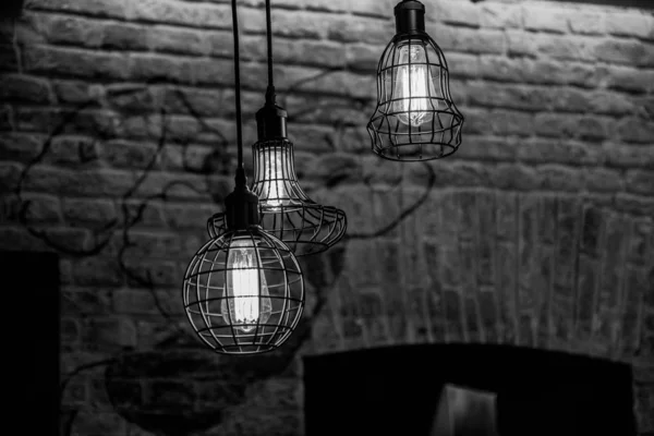 Black and white photography of wire lamps hanged from ceiling on old brick wall background. Glowing filaments inside glass lightbulbs. Geometric carcass shapes. Urban style interior lighting concept. Wire chandelier closeup. Geometric wire carcasses