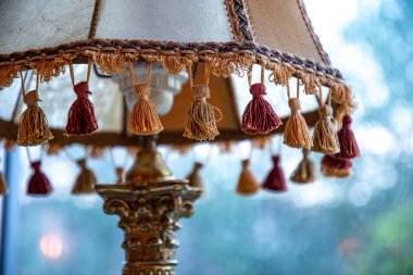 Closeup of brown tassels on antique lampshade with ornate bronze foot lamp element and blurry background. Vintage lamp details. Old-fashioned interior object. Home decor item. Rustic style lighting. Cozy grannie's home. clipart