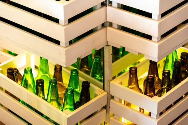 Zigzag shaped shelves made from white painted wood crates with green and brown glass bottles inside. Retro style beer bottle rows. Empty glass bottles shined by lighting. Interior decorations with reused wood crates and reused empty glass bottles.