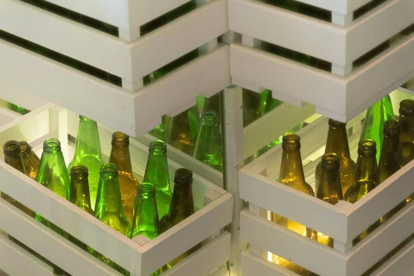 Corners of shelves made from white painted wood crates with transparent glass bottles inside. Retro style beer bottle rows. Empty green and brown glass bottles shined by interior lighting. Interior decoration with reused wood crates and empty bottles