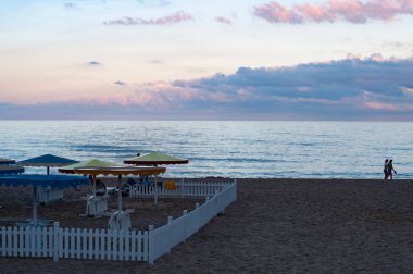 Çift siluet açık mavi sakin deniz suyuna karşı yürüyor. Alacakaranlık akşamplaj şemsiye siluetleri ile Yaz deniz manzarası. Koyu plaj kumu üzerinde beyaz ahşap çit. Karadeniz sahili seyahat 