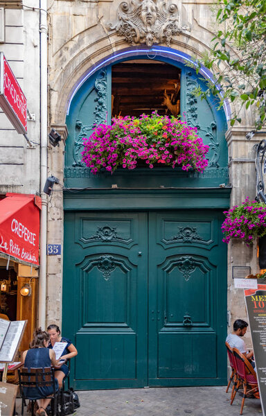 Париж / Франция - 06 26 2019: Красивая декоративная бирюзовая дверь с фиолетовыми цветами и людьми, сидящими в уличном кафе. Архитектура барокко Парижа. Детали внешней отделки античного здания
.