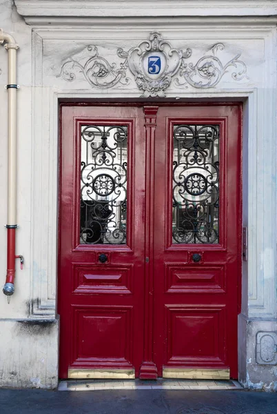 Antique red door with framed wooden panels and ornate grids on door windows of old building in Paris France. Vintage doorway and drain pipe on fretwork stone wall of house in baroque architecture.