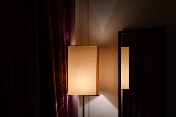 Geometry of simple lampshade reflected in mirror on wall of room with dark red ornate curtains. Golden light and triangle shadows in corner of room in darkness. Minimal interior decoration.