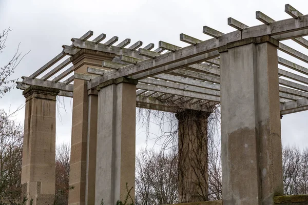 Classical architecture style arbor in Tiergarten park of Berlin Germany. Aged stone columns of ancient arbor with striped wooden roof. Garden house overgrown with bare vine in winter season.