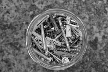 Çeşit çeşit donanım cıvataları ve cam kavanozdaki fındıklar ile grunge taşı arka planı ve kopyalama alanı içeren siyah beyaz fotoğraf. Cam kavanozun içindeki metal eşya deposu