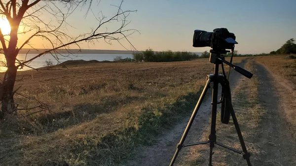 三脚架上的专业摄影师相机站在光秃秃的树旁的土路上 望着落日洒落在水面上 乡间风景 有架着相机 没有人 — 图库照片