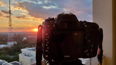 Odessa Ukrayna - 08 27 2020: Tripod üzerindeki DSLR kamerası bulutlu gökyüzü ve iletişim kulesinin bulanık silueti ile gün batımını çekiyor