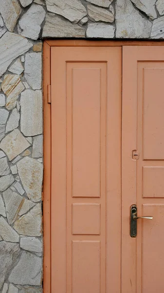 Panels of wooden door painted in light orange color and rough stone wall texture nearby. Exterior of old building with retro double door and stone facing wall