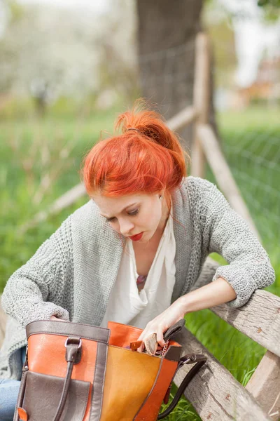 彼女の財布の中に何かを探して外のベンチに座っている赤い髪の美しい若い女性のハンドバッグ — ストック写真