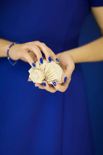 ブルーのドレスの前に小さな海カタツムリの殻を持って完璧なマニキュアと美しい女性の手をバック グラウンドとして使用できます — ストック写真